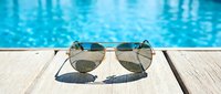 Pair of aviator sunglasses with mirrored lenses resting on wooden poolside deck, reflecting sparkling blue water, summer vacation and leisure concept.