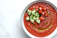 Chilled tomato gazpacho soup garnished with diced cucumber and tomatoes in white bowl, drizzled with olive oil on marble surface. Fresh summer appetizer.