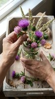 Close-up of mature hands arranging purple thistle flowers and dried wheat stems in a glass vase on a wooden tray by a window, capturing a moment of floral arrangement and home decoration.