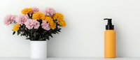Fresh pink and yellow chrysanthemums in white ceramic vase next to orange cosmetic dispenser bottle on white shelf against light wall, minimal home decor.