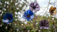 Colorful glass flower decorations hanging on delicate wire branches against soft blurred natural background. Handcrafted art pieces for garden decor.