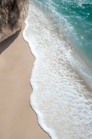 Aerial view of turquoise ocean waves washing up on sandy beach with dramatic coastal cliff, creating natural curves where water meets pristine shore.
