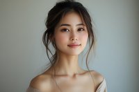 Young Asian woman with natural makeup and loose updo hairstyle smiling softly against neutral background, bare shoulders creating elegant portrait mood.