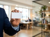Close-up of male hand holding blank white business card in modern office environment with colleagues working in background.