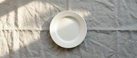 Empty white ceramic plate on wrinkled linen fabric with natural sunlight shadows creating minimalist tableware composition for food photography and design.