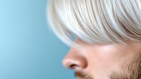 Close up profile portrait of young Caucasian male with platinum blonde hair against light blue background, showing partial face and modern hairstyle.