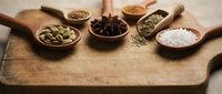 Aromatic spices and seasonings arranged in wooden spoons on rustic cutting board for culinary cooking and food preparation in kitchen.