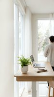 Modern home office workspace with young man standing by large windows overlooking green trees. Minimalist desk setup with laptop and plant for remote work.