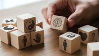Hand placing wooden cube with brain symbol among blocks featuring yin yang, human figures and question marks on wood surface for concept design.