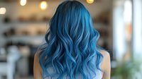 Young woman with vibrant blue wavy ombre hair styled in loose curls, viewed from behind in salon setting with soft bokeh lights and blurred background.