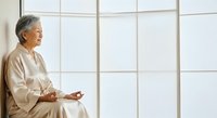 Senior Asian woman in beige linen shirt sitting by large window, meditating with hands in prayer position, peaceful mindfulness and wellness concept for relaxation and mental health.