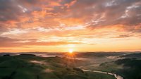 Aerial view of rolling green hills with winding river at golden hour sunrise, dramatic orange and pink sky with clouds, misty valley landscape for nature and travel projects.