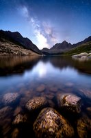 Serene mountain lake reflecting starry night sky with Milky Way galaxy above rugged peaks, clear water revealing stones beneath.