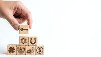 Hand placing wooden cube with key symbol on pyramid of blocks with luck symbols including horseshoe, clover, sun and dollar sign on white background.