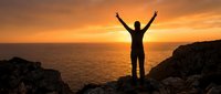 Silhouette of person with arms raised celebrating success on rocky cliff overlooking ocean at golden sunset, representing achievement, freedom, and triumph.
