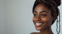 Young African American woman with radiant smile wearing black and gold head wrap against light background, natural makeup look and glowing skin for beauty and lifestyle content.