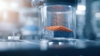 Orange powder particles floating in transparent laboratory container against blurred scientific background, demonstrating molecular behavior in controlled environment.