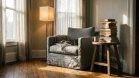Cozy reading nook with upholstered armchair, stacked books on wooden side table, floor lamp, and sheer curtains in warm, natural light for interior design and home decor projects.