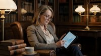 Middle-aged woman with light brown hair and glasses, wearing a tan blazer, sitting in a leather armchair in a classic library setting, holding and viewing a digital tablet, surrounded by books, warm desk lamps, and wooden bookshelves, representing professional work, research, and modern technology in traditional environments.