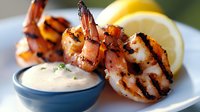 Grilled jumbo shrimp with charred grill marks served alongside creamy dipping sauce and fresh lemon wedges on white plate, macro food photography.