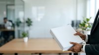 Businessman holding blank white box in modern office workspace with colleagues and plants in background.