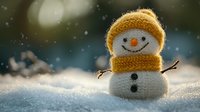 Adorable knitted snowman with orange hat and scarf sits in fresh snow during gentle snowfall, creating cozy winter holiday atmosphere for seasonal decorations.