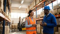 Two diverse warehouse workers in safety gear discussing logistics operations in modern distribution center with shelving and forklift background.