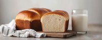 Fresh baked white bread loaf sliced on wooden cutting board with glass of milk and kitchen towel, soft morning light, homestyle food photography.