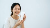Young Asian woman with dark hair smiling while holding white skincare bottle against neutral background for cosmetics and beauty product marketing.
