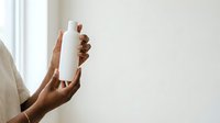 Woman with dark skin tone holding blank white cosmetic bottle against neutral background, suitable for beauty product packaging mockups and skincare advertising campaigns.