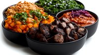 Assorted bowls with grilled meat, roasted vegetables, fresh herbs, and coleslaw served in modern black ceramic dishes on white background, ready for food service.
