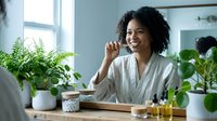 Young African American woman in bathrobe enjoying morning coffee at kitchen table surrounded by green plants creating peaceful wellness lifestyle moment.