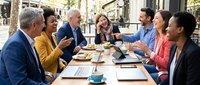Diverse group of business professionals dining outdoors at a table, engaged in conversation and meal during lunch break or corporate meeting in urban setting.