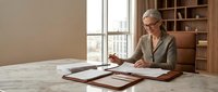 Professional mature Caucasian businesswoman with glasses reviewing documents at modern office desk near window with city view for corporate presentations.