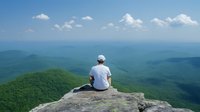 Caucasian male in white shirt sits alone on mountain cliff edge overlooking vast green forest landscape under blue summer sky with white puffy clouds.