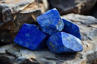 Raw lapis lazuli stones with deep blue color and golden pyrite inclusions displayed on dark rock, showing natural crystalline structure and mineral texture.