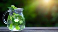 Fresh cucumber mint infused water in glass pitcher on wooden surface against blurred green garden background, refreshing summer drink detox.