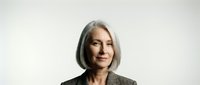 Mature woman with long gray hair and olive complexion wearing dark blazer against neutral background, suitable for corporate, business, and professional portraits.