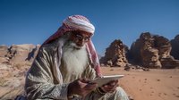 Senior Middle Eastern man with white beard wearing traditional headscarf using digital tablet in desert landscape with rocky mountains against clear blue sky.
