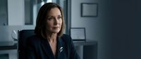 Middle-aged woman with long dark hair and fair complexion, wearing professional dark blazer with pin, seated in modern office environment with blurred background.