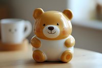 Cute ceramic teddy bear figurine with honey-colored head and paws, white body, and smiling face on wooden table with blurred cup in background.