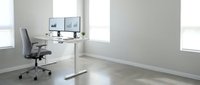 Modern minimalist home office workspace with height-adjustable white desk, ergonomic gray chair, dual monitor setup in bright room with large windows and polished floors.