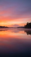 Serene lake at sunrise with vibrant orange and purple sky reflecting on calm water, surrounded by forested shoreline and morning mist for nature and landscape projects.