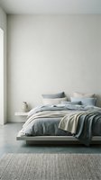 Minimalist bedroom interior with low platform bed, layered bedding in neutral tones, decorative pillows, ceramic vase, and woven area rug against white walls.