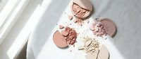Broken makeup powder compacts in nude and pink shades scattered on white surface with natural window light creating soft shadows for beauty photography.