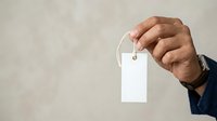 Male hand holding blank white price tag or label with string against neutral background for retail, product pricing, or promotional messaging.