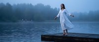 Young woman with long brown hair and light skin tone wearing flowing white dress standing on wooden dock extending arms toward misty lake surrounded by evergreen forest at dawn or dusk.
