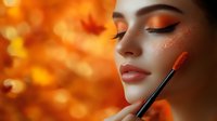 Young woman with autumn-inspired makeup and glitter details against blurred orange bokeh background with maple leaves, makeup brush applying blush to cheeks.