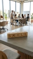 Wooden blocks spelling skill on modern office desk with diverse team meeting in bright corporate workspace with large windows and plants.