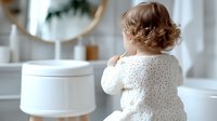 Young Caucasian toddler in white polka dot dress stands at bathroom counter reaching up, back view in bright modern minimalist interior with round mirror.
